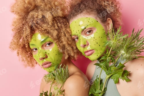 Preview: Beauty and natural cosmetics concept. Curly haired sisters apply green moisturising masks on face ho