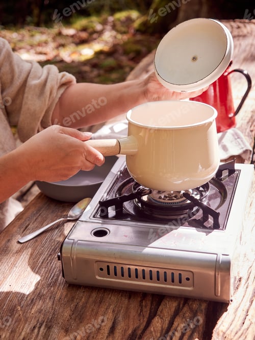 Preview: hand opening the lid of a pot placed on a portable gas stove on a wooden table, cooking outdoors.