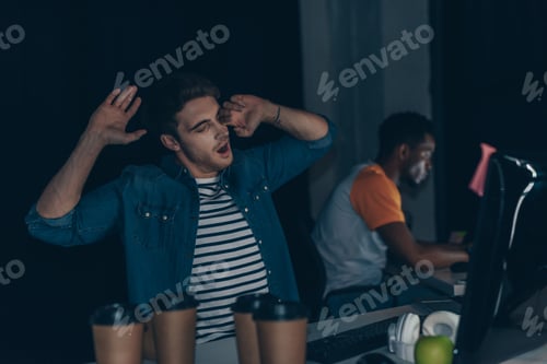 Preview: selective focus of young programmer yawning while sitting near working african american colleague