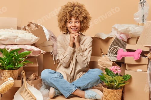 Preview: Positive thoughtful curly woman keeps hands under chin sits crossed legs on floor with packed carton