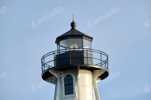 Preview: White tall lighthouse on sea shore against blue sky for commercial vessels navigation