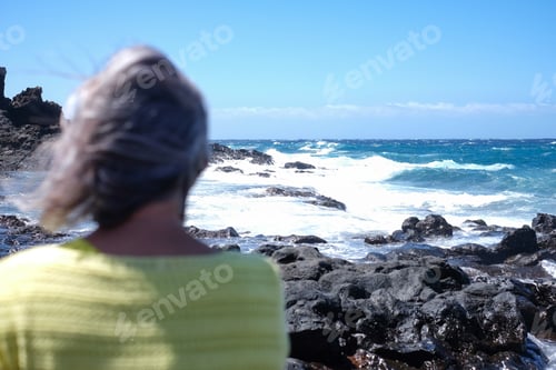 Preview: Rear view of defocused gray haired woman on a rocky beach looking at ocean waves crash on cliffs