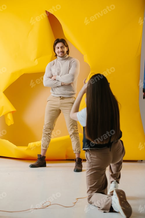 Preview: Female photographer taking pictures of professional male model on torn paper background in studio.