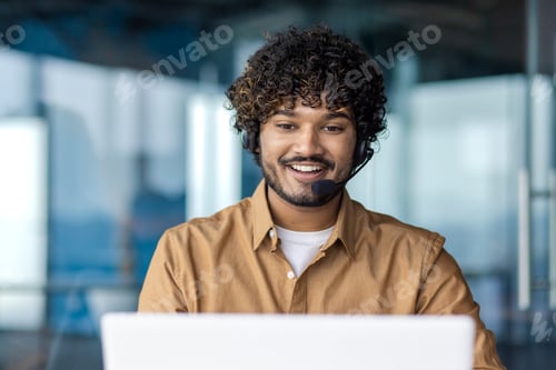 Preview: Smiling arab man with curly hair in wireless headset looking at laptop screen while sitting in