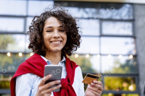 Preview: Beautiful smiling woman holding mobile phone and credit card buying looking at camera on the street