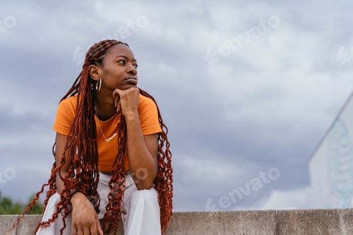 Preview: Pensive young woman with braids sitting under cloudy sky