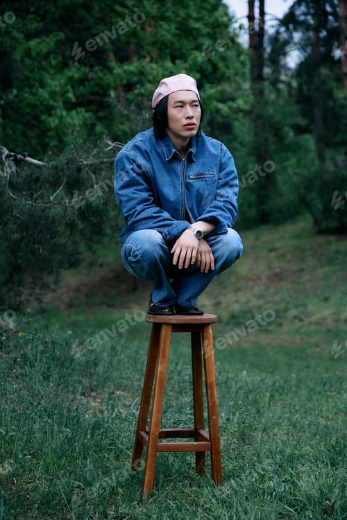 Preview: Young man in stylish denim outfit poses creatively in a serene park setting