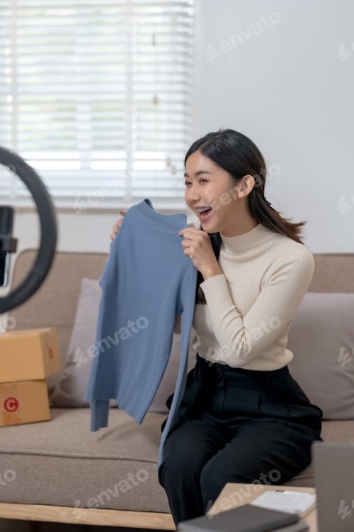 Preview: Smiling woman showcasing a blue sweater for an online audience with a smartphone and ring light.
