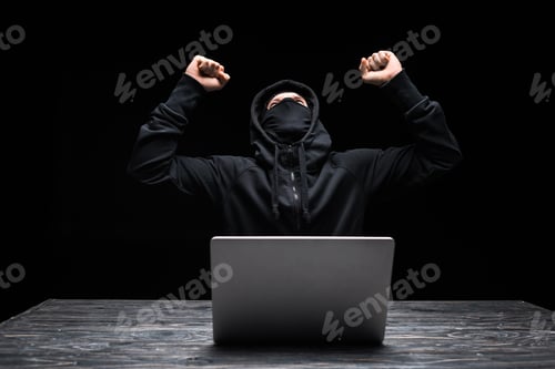 Preview: excited hacker in mask celebrating triumph near laptop isolated on black