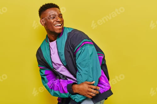 Preview: excited african american man in bright windbreaker jacket laughing and looking away on yellow