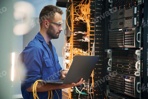 Preview: laptop in hands. Young man is working with internet equipment and wires in server room