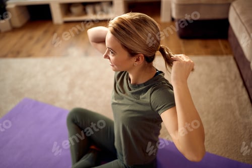 Preview: Happy athletic woman tying her hair in ponytail while getting ready for home workout.