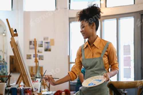 Preview: Creative young woman wearing apron painting picture with brush on canvas in art studio.