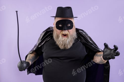 Preview: Mature bearded man with overweight wearing Zorro suit with toy epee poses on purple background