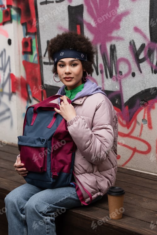 Preview: African american woman holding backpack near paper cup on bench and graffiti