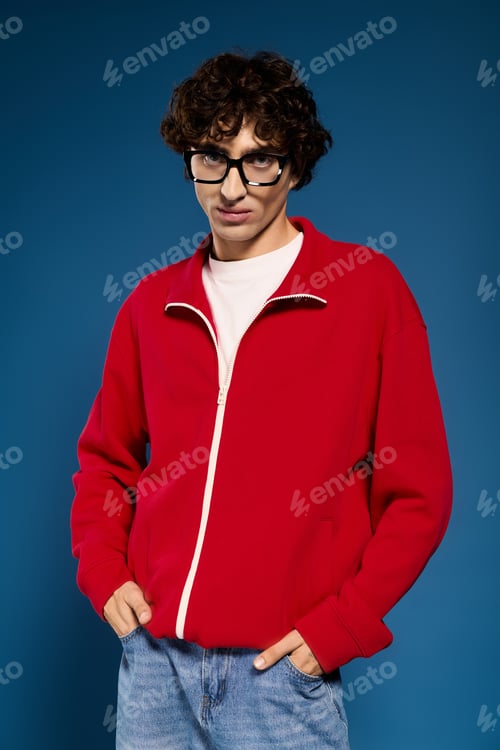 Preview: Stylish young man showcases vibrant red sweater against blue backdrop in trendy attire