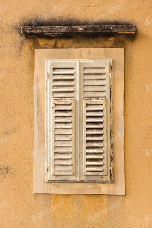 Preview: Closed window with white shutters on the yellow wall of an old house