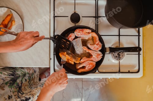 Preview: Hands with fork and skimmer frying salmon fish in hot pan on gas stove at home