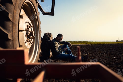 Preview: Farmer siting and using tablet while resting after cultivation of soil leaning on tractor tire.