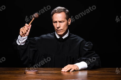 Preview: judge in judicial robe sitting at table and hitting with gavel isolated on black