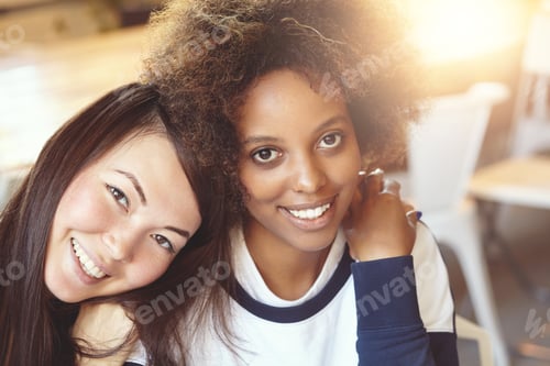 Preview: Portrait of two beautiful women of different ethnicities sitting at cafe, hugging and having fun. At