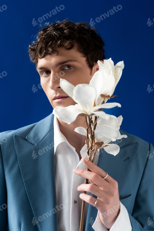 Preview: Trendy young man in jacket and shirt holding magnolia flowers isolated on blue