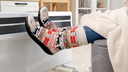 Preview: Closeup of woman wearing warm socks sitting at electric heater in living room