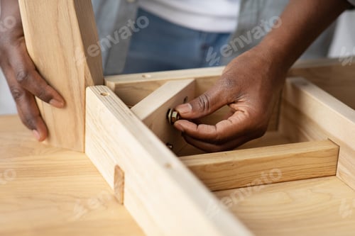 Preview: Self-assembly furniture concept. Black man screwing bolt to wooden table, installing desk by