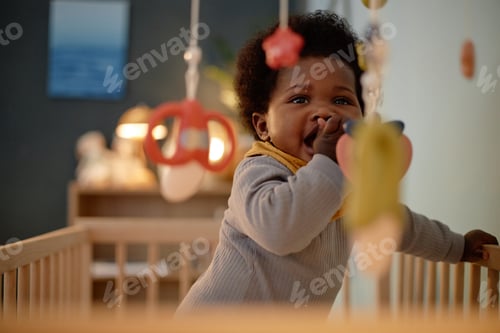 Vista previa: Happy Black Baby Girl Biting Teether while Standing in Crib in Bedroom