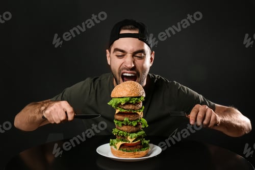 Preview: Young hungry man with cutlery eating huge burger on black background