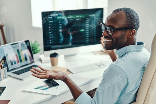 Preview: Handsome young African man in shirt using computer and smiling while working in the office