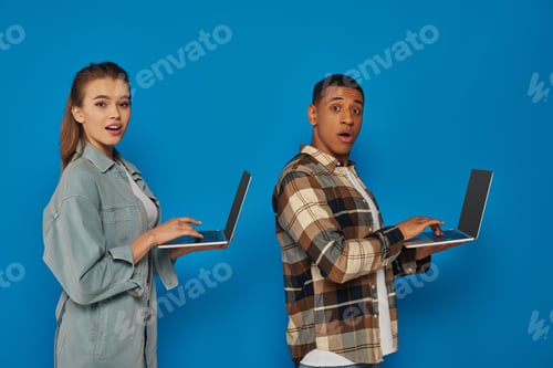 Preview: amazed interracial freelancers using laptops on blue backdrop, diverse cultures man and woman