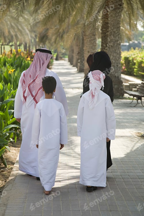 Preview: A family dressed in traditional Arabic clothes walking down a palm treelined pathway.