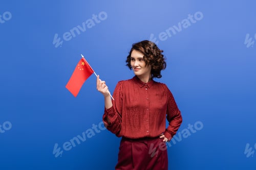 Preview: happy language teacher holding flag of China while standing with hand on hip isolated on blue