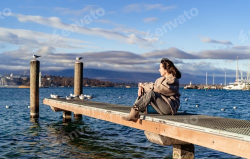 Preview: Happy carefree woman sitting on pier on the lake with seagulls enjoying fresh autumn breeze