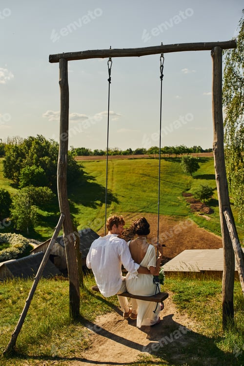 Preview: rustic wedding, back view of young newlyweds on swing in countryside with green scenic landscape