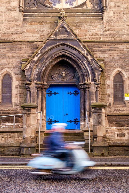 Preview: Person on a scooter driving past a gothic arch with bright blue door.