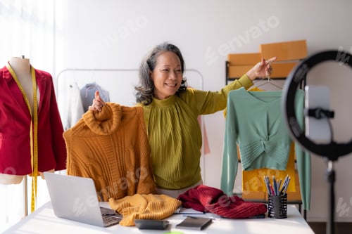 Preview: Asian senior female online seller reviewing shirt video live streaming to social media at home.