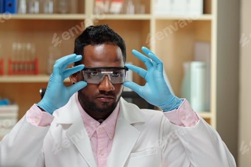 Preview: Young black man in lab coat and gloves putting on protective eyeglasses