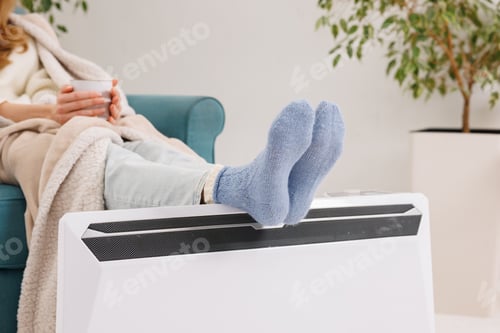 Preview: Young girl in warm knitted socks warms her feet on a radiator. Heating season concept