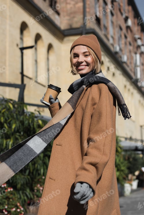Preview: joyful young woman in beige coat and hat holding paper cup with coffee to go