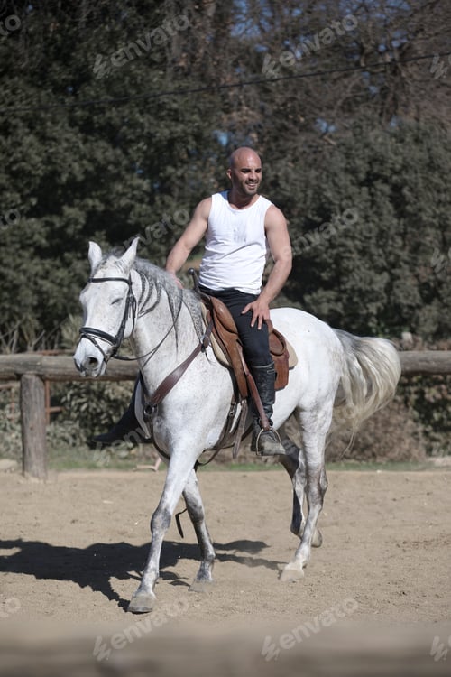 Preview: Young guy in casual outfit riding white horse on sandy ground
