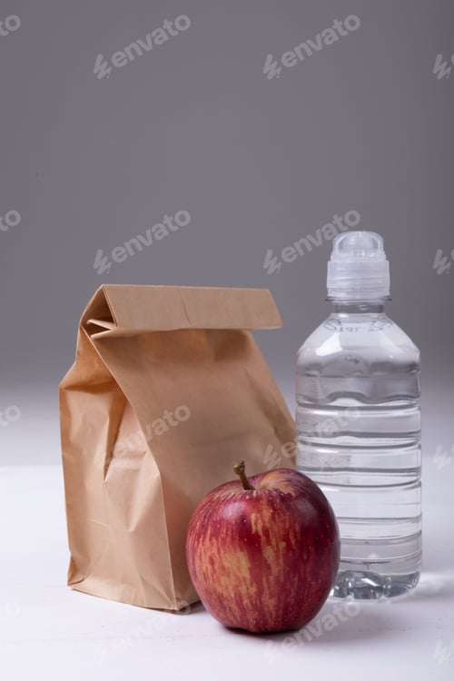 Preview: Close-up of brown paper lunch bag with apple and water bottle on table against gray background