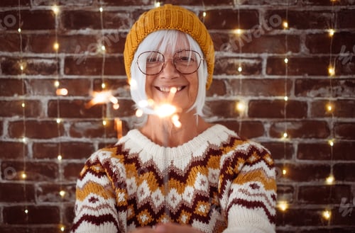 Preview: Handsome senior caucasian woman with eyeglasses and winter sweater holding sparkler light