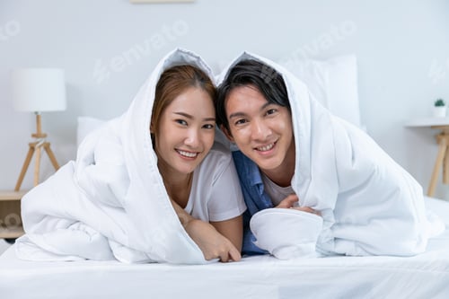 Preview: Couple playing in bed at home, hugging soft pillow, nose touching relaxed at home on bed