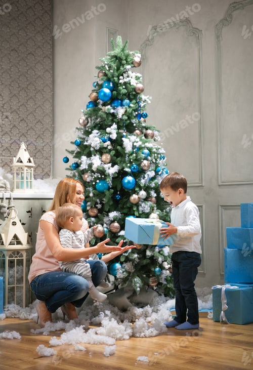 Preview: Young mother giving a present to the oldest son, baby boy touching bauble on Christmas tree