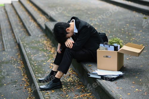 Preview: Upset businessman sitting hopelessly on stairs of building with box of personal stuff.