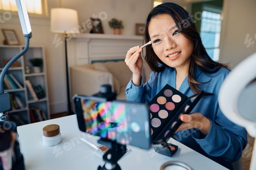 Preview: Happy Asian woman eyeshadow palette while recording makeup tutorial at home.
