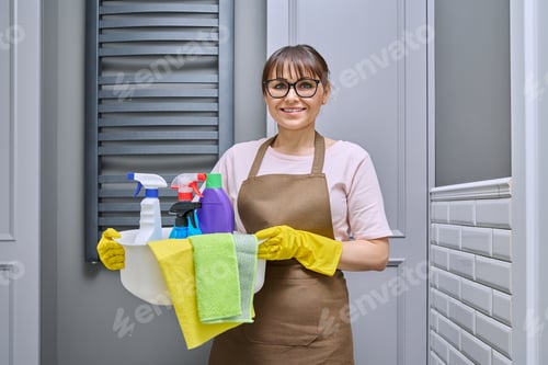 Preview: Middle-aged woman in gloves apron with basin of detergents in bathroom