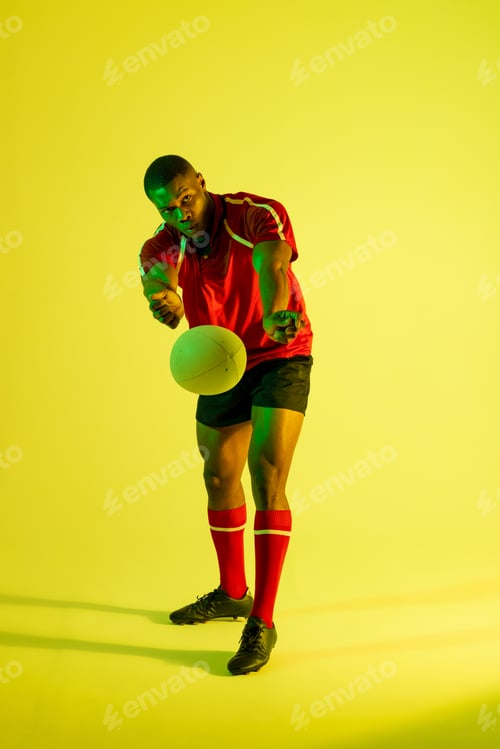 Preview: African american male rugby player with rugby ball over yellow lighting